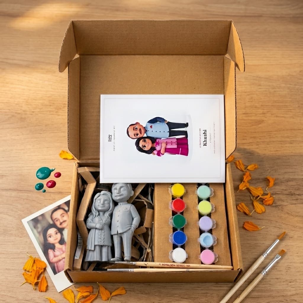 An open cardboard kit box on a wooden table showing two grey unpainted figurines, a printed reference photo, rows of small paint pots, and two brushes, with marigold petals scattered around
