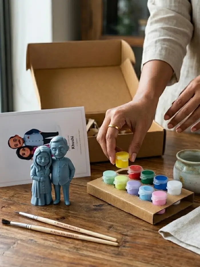 A hand reaching for a yellow paint pot in a kit of small colour pots on a wooden table, next to an unpainted grey figurine of a couple, a guide card, two brushes, and an open kraft box